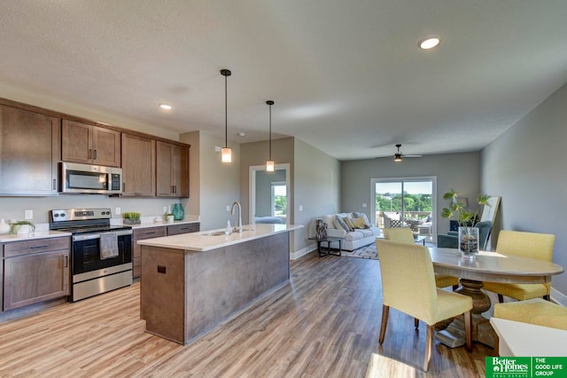 kitchen with light hardwood / wood-style floors, appliances with stainless steel finishes, an island with sink, decorative light fixtures, and ceiling fan