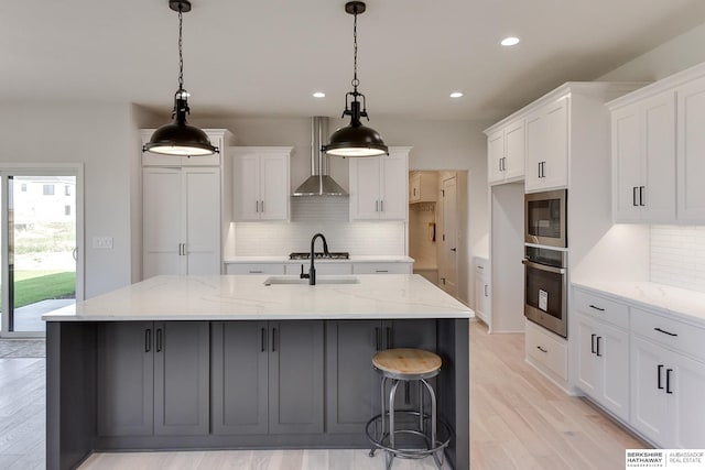 kitchen with white cabinets, a center island with sink, light hardwood / wood-style flooring, wall chimney range hood, and appliances with stainless steel finishes