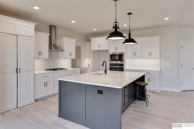 kitchen with a kitchen island with sink, light hardwood / wood-style floors, pendant lighting, wall chimney range hood, and white cabinetry