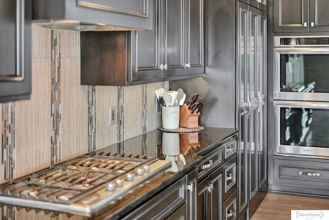 kitchen with dark brown cabinets, ventilation hood, stainless steel appliances, light hardwood / wood-style flooring, and dark stone countertops