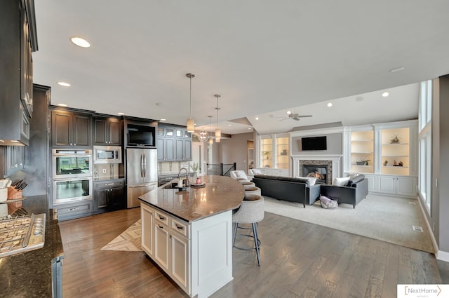 kitchen featuring pendant lighting, a breakfast bar, a kitchen island with sink, built in shelves, and appliances with stainless steel finishes