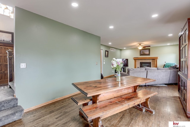 dining space with a tiled fireplace, ceiling fan, and dark wood-type flooring