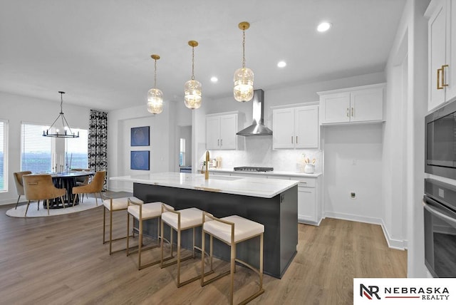 kitchen with light hardwood / wood-style floors, stainless steel appliances, an island with sink, wall chimney exhaust hood, and pendant lighting