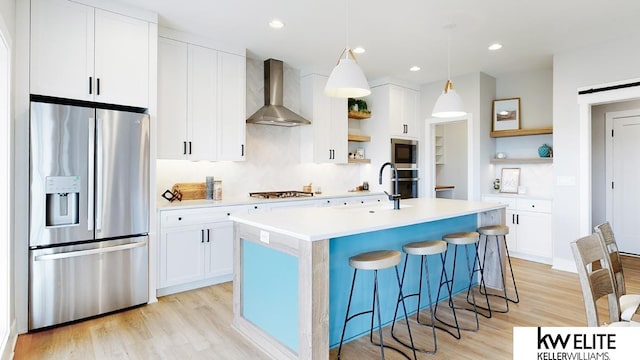 kitchen featuring stainless steel appliances, pendant lighting, a kitchen island with sink, light wood-type flooring, and wall chimney exhaust hood