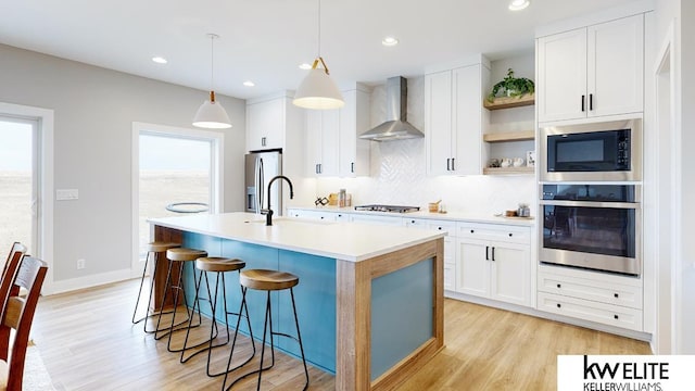 kitchen featuring light hardwood / wood-style floors, appliances with stainless steel finishes, a center island with sink, and wall chimney exhaust hood