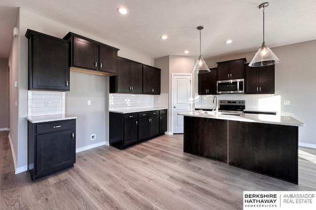 kitchen featuring stainless steel appliances, tasteful backsplash, a kitchen island with sink, hanging light fixtures, and light hardwood / wood-style flooring