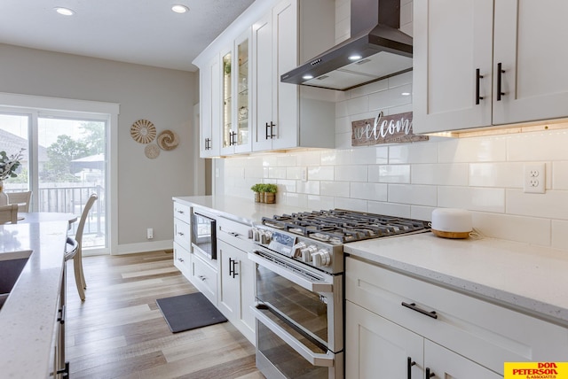 kitchen featuring light wood-type flooring, white cabinets, backsplash, wall chimney exhaust hood, and range with two ovens