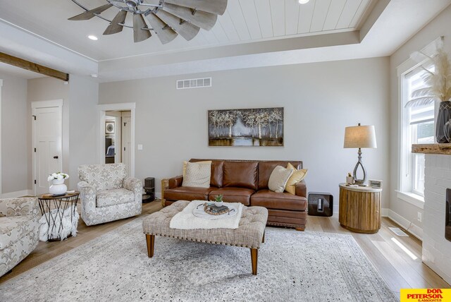 living room featuring ceiling fan, a tray ceiling, light wood-type flooring, and a brick fireplace