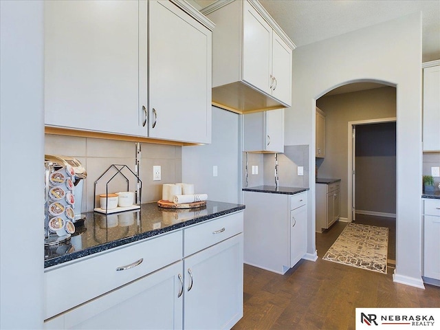 kitchen featuring arched walkways, white cabinetry, dark stone countertops, dark wood-style flooring, and decorative backsplash