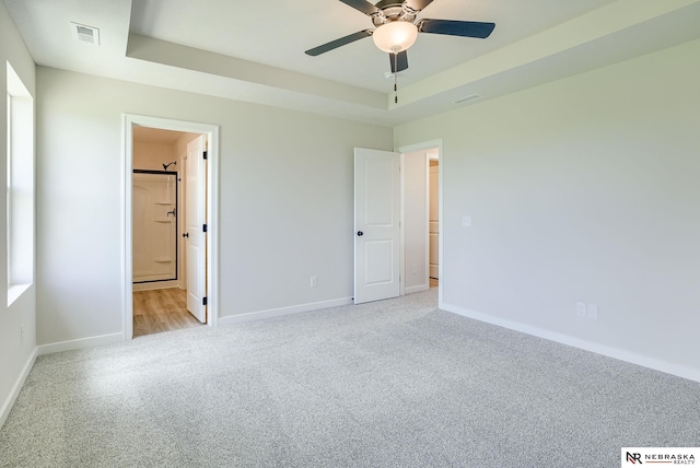 unfurnished bedroom with ceiling fan, a tray ceiling, connected bathroom, and light carpet