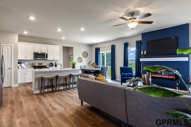 living area featuring baseboards, a ceiling fan, a glass covered fireplace, light wood-type flooring, and recessed lighting