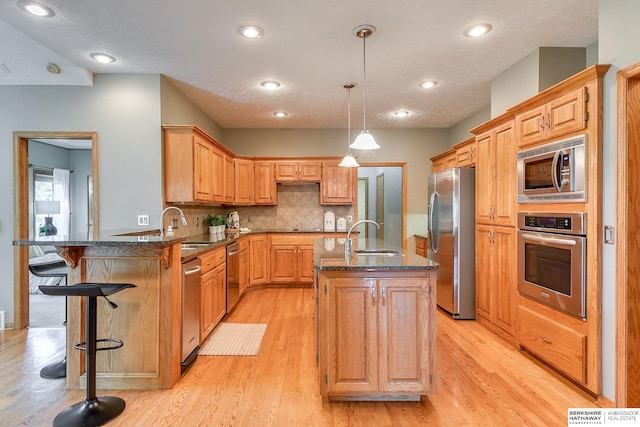 kitchen with decorative light fixtures, a center island with sink, light hardwood / wood-style flooring, appliances with stainless steel finishes, and decorative backsplash