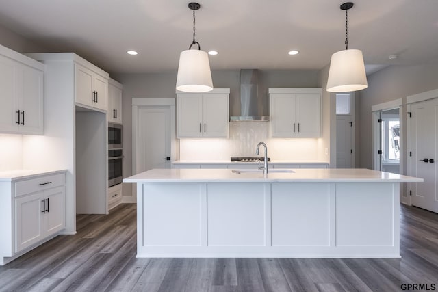 kitchen with wall chimney range hood, a kitchen island with sink, white cabinets, and a sink