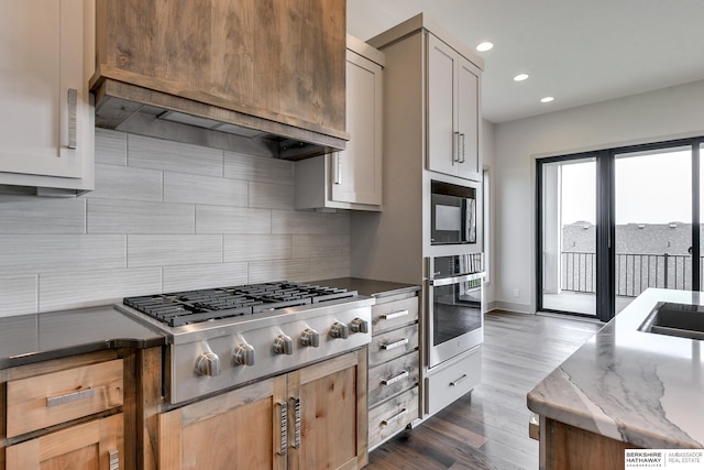 kitchen with stainless steel gas cooktop, backsplash, custom range hood, dark stone countertops, and dark hardwood / wood-style flooring