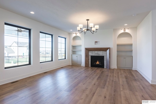 unfurnished living room featuring built in shelves, ceiling fan with notable chandelier, light hardwood / wood-style floors, and a high end fireplace