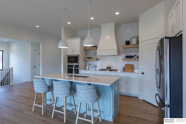 kitchen featuring appliances with stainless steel finishes, an island with sink, white cabinets, light wood-type flooring, and decorative light fixtures