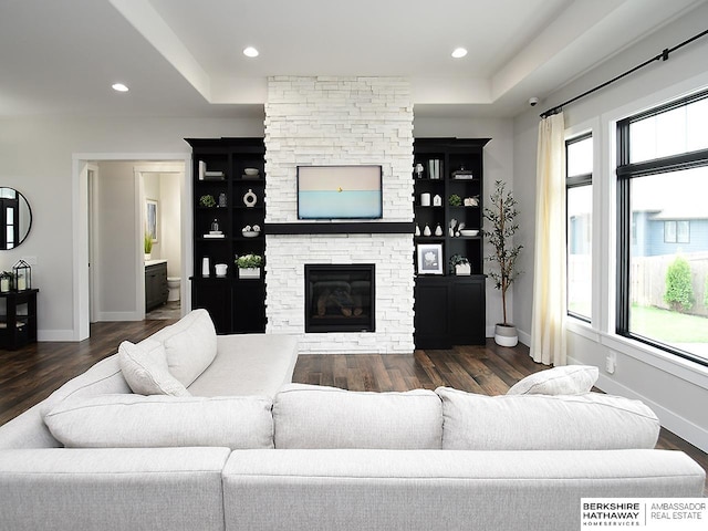 living room with a stone fireplace, built in shelves, a tray ceiling, and dark hardwood / wood-style flooring