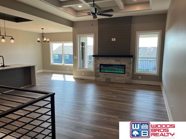 unfurnished living room with coffered ceiling, dark wood-type flooring, ceiling fan with notable chandelier, a stone fireplace, and a wealth of natural light