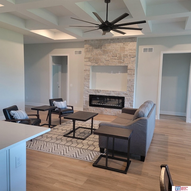 living room featuring ceiling fan, a stone fireplace, light wood-type flooring, and coffered ceiling