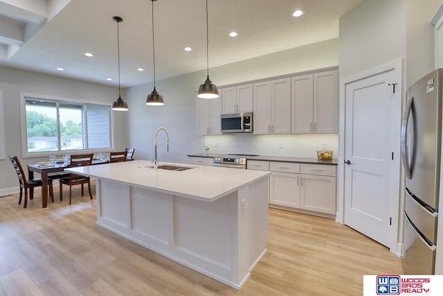 kitchen with hanging light fixtures, sink, a center island with sink, stainless steel appliances, and light wood-type flooring