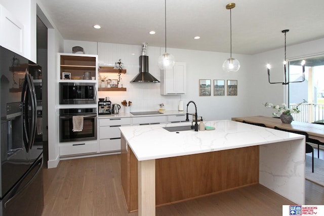 kitchen featuring a kitchen island with sink, white cabinets, wall chimney exhaust hood, black appliances, and light stone countertops