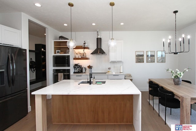 kitchen with light stone counters, white cabinets, wall chimney exhaust hood, black appliances, and a center island with sink