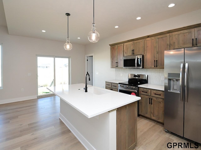 kitchen with appliances with stainless steel finishes, an island with sink, light wood-type flooring, decorative light fixtures, and sink