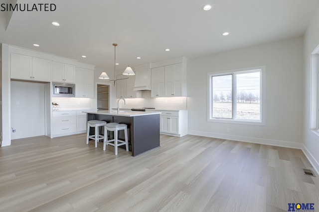 kitchen with light wood-type flooring, an island with sink, white cabinetry, stainless steel microwave, and decorative light fixtures