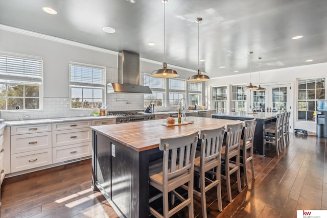 kitchen featuring wall chimney exhaust hood, white cabinets, a kitchen island with sink, and decorative light fixtures