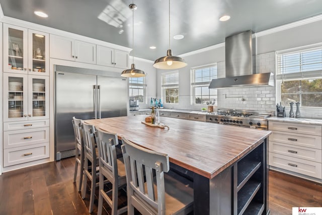 kitchen with wall chimney range hood, hanging light fixtures, a kitchen island, appliances with stainless steel finishes, and white cabinetry