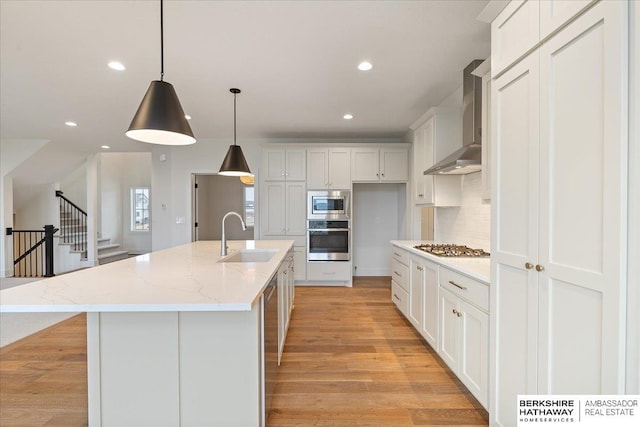 kitchen with appliances with stainless steel finishes, white cabinetry, sink, a center island with sink, and wall chimney range hood