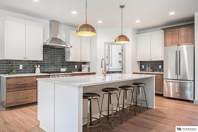 kitchen featuring wall chimney range hood, a kitchen island with sink, light wood-type flooring, stainless steel appliances, and white cabinets