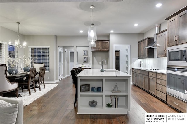 kitchen with dark brown cabinets, stainless steel appliances, dark wood-type flooring, decorative light fixtures, and an island with sink