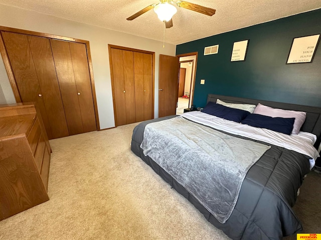 bedroom with a textured ceiling, two closets, light colored carpet, and ceiling fan