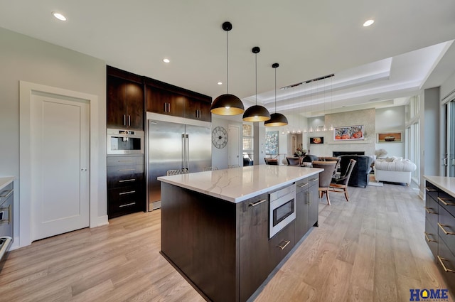 kitchen featuring a kitchen island, light hardwood / wood-style flooring, built in appliances, dark brown cabinetry, and a fireplace