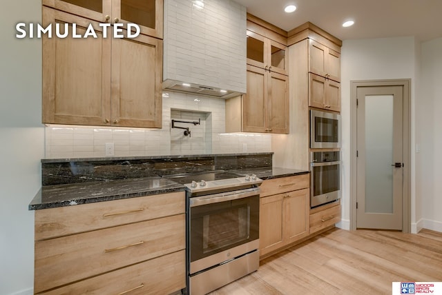 kitchen featuring wall chimney exhaust hood, light hardwood / wood-style flooring, stainless steel appliances, dark stone countertops, and light brown cabinetry