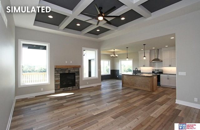 kitchen with stainless steel range with electric cooktop, a kitchen island with sink, wall chimney exhaust hood, decorative light fixtures, and white cabinetry