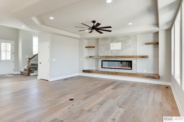 unfurnished living room featuring a tiled fireplace, a tray ceiling, ceiling fan, and light wood-type flooring