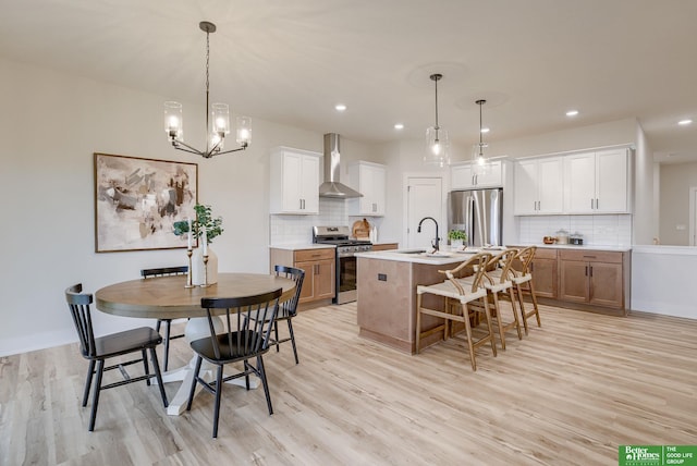 kitchen with appliances with stainless steel finishes, an island with sink, white cabinetry, light hardwood / wood-style floors, and wall chimney exhaust hood