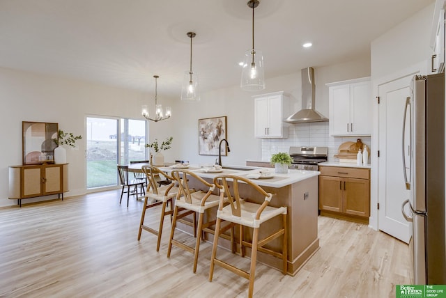 kitchen with wall chimney range hood, appliances with stainless steel finishes, an island with sink, hanging light fixtures, and white cabinets