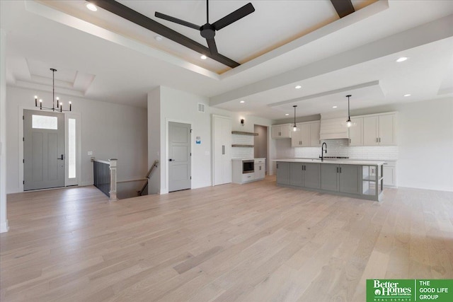 unfurnished living room with light hardwood / wood-style floors, ceiling fan with notable chandelier, sink, and a tray ceiling