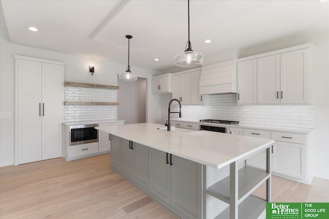 kitchen featuring white cabinets, an island with sink, pendant lighting, light hardwood / wood-style flooring, and premium range hood