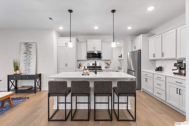 kitchen with appliances with stainless steel finishes, decorative light fixtures, a kitchen island with sink, white cabinets, and light wood-type flooring