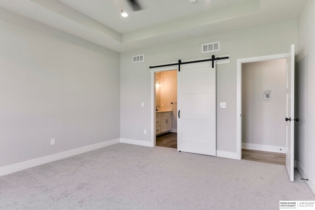 unfurnished bedroom featuring a barn door, ensuite bathroom, light carpet, and a tray ceiling