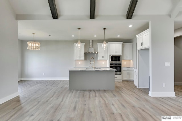 kitchen featuring appliances with stainless steel finishes, beam ceiling, pendant lighting, wall chimney exhaust hood, and a kitchen island with sink