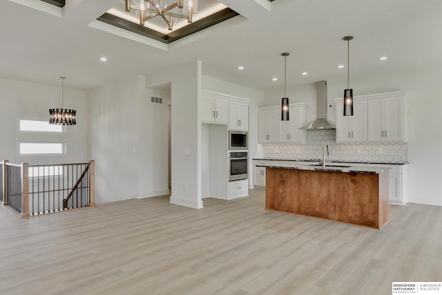 kitchen with stainless steel appliances, light hardwood / wood-style floors, white cabinets, wall chimney exhaust hood, and an island with sink