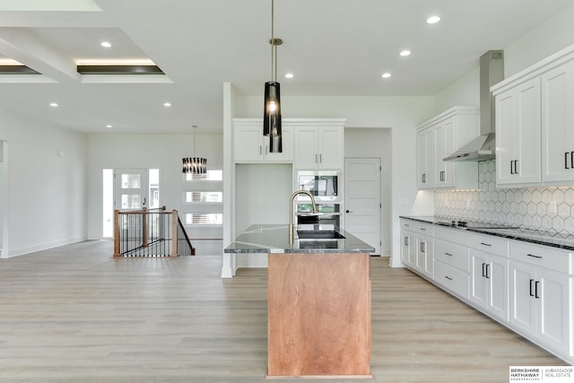 kitchen with an island with sink, light hardwood / wood-style floors, wall chimney exhaust hood, and dark stone countertops