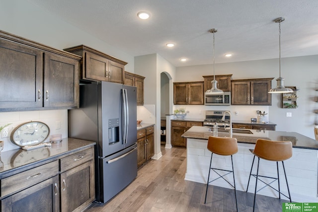 kitchen with light hardwood / wood-style floors, appliances with stainless steel finishes, a kitchen breakfast bar, decorative light fixtures, and an island with sink