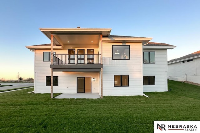 back house at dusk with a patio, a balcony, and a lawn