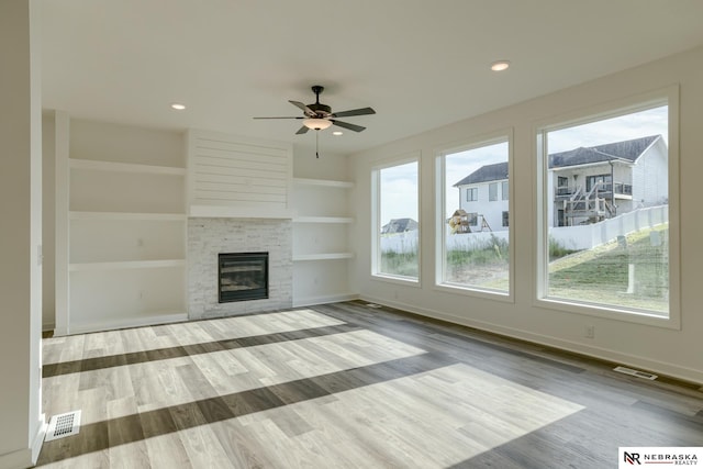 unfurnished living room with built in shelves, ceiling fan, a stone fireplace, and light wood-type flooring